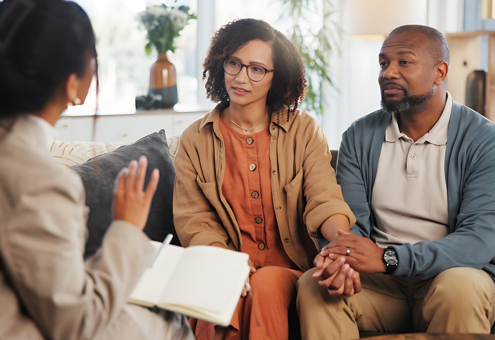 A couple sits on a couch, holding hands, while talking to a person with a notebook. The setting appears to be a well-lit office or therapy room.