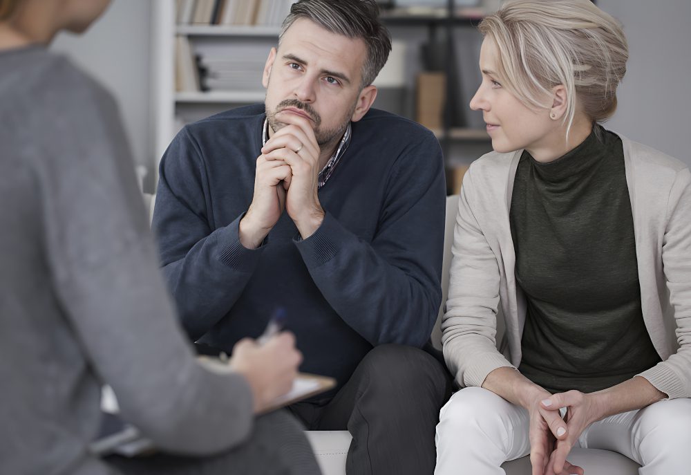 A man and woman sit closely together, attentively listening to a person speaking out of frame. They are in a room with bookshelves in the background.
