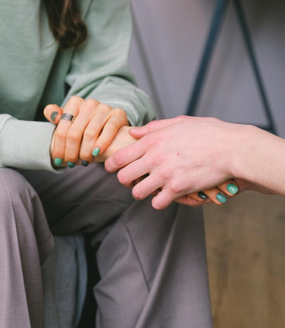 Two people sitting close, holding hands. Both have teal nail polish and light skin. They are wearing casual clothing in shades of green and gray.
