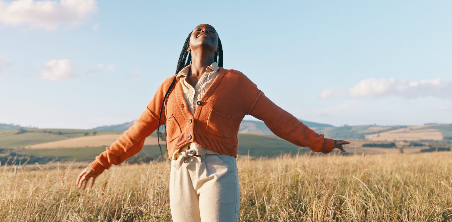 Person standing in a grassy field with arms outstretched, wearing an orange cardigan and beige pants, under a blue sky with scattered clouds.