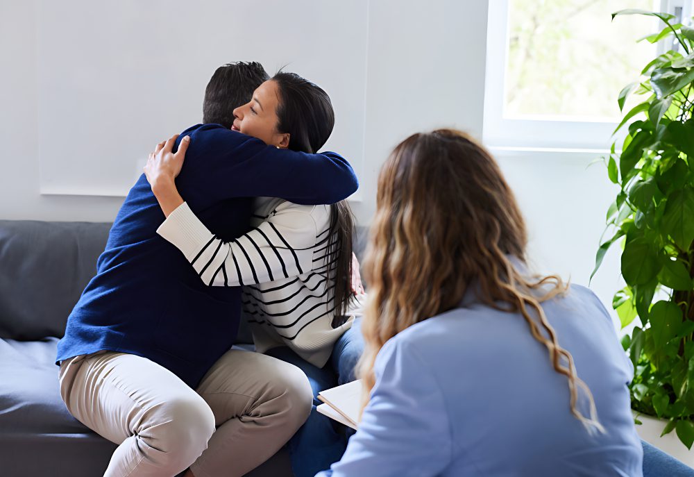 Two people are hugging on a couch in a therapist's office while a third person, possibly a therapist, observes and takes notes. There is a plant next to the window in the background.