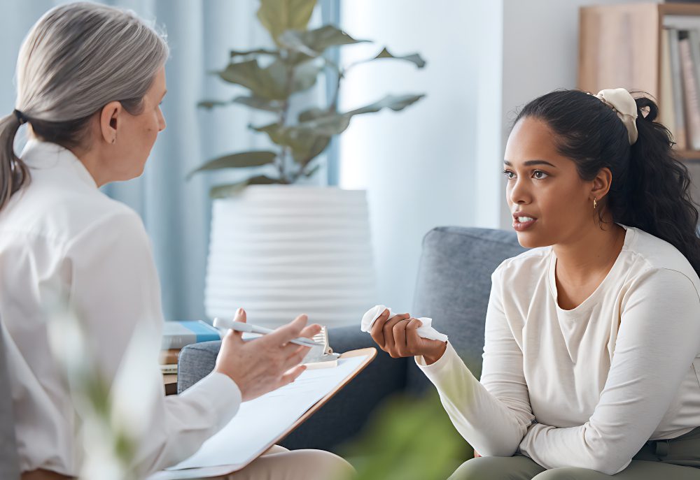 A woman with a ponytail sits on a couch holding a tissue, speaking to another woman with gray hair who is taking notes on a clipboard. They appear to be having a serious conversation.