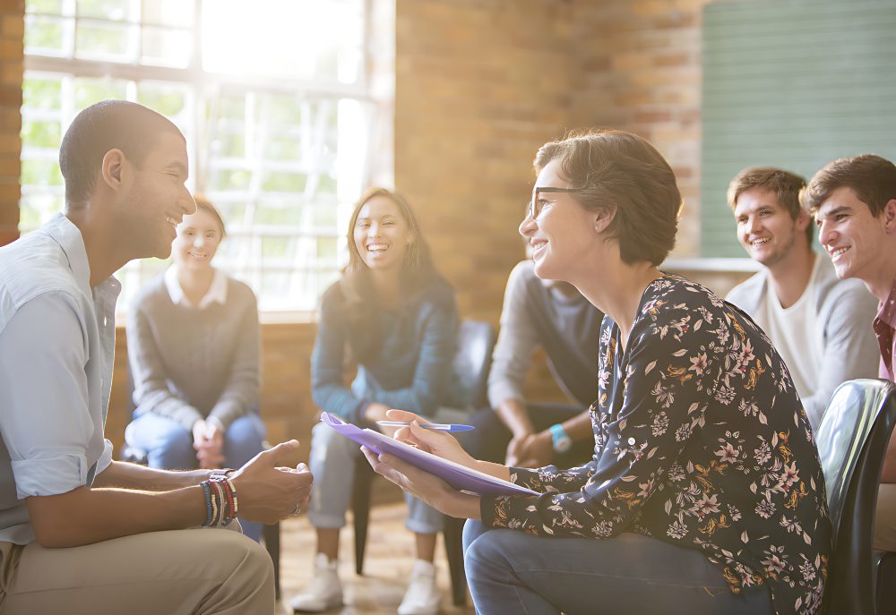 A group of people sitting in a circle, engaged in discussion. A woman with glasses writes on a clipboard, while others listen and smile. A bright window is in the background.