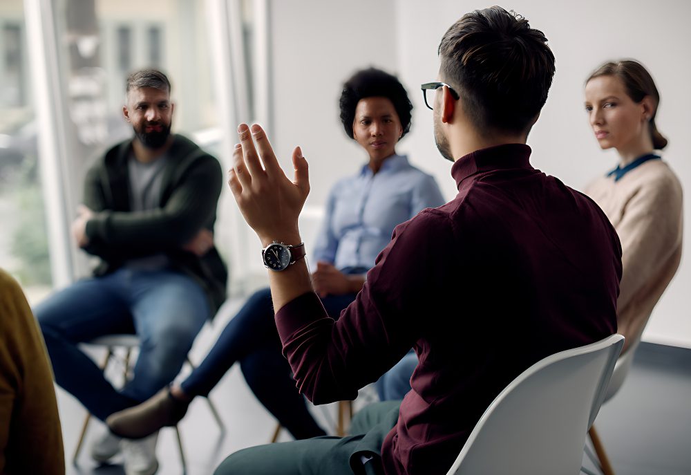 A group of people sitting in a circle, engaged in discussion. The person in the foreground gestures while speaking.